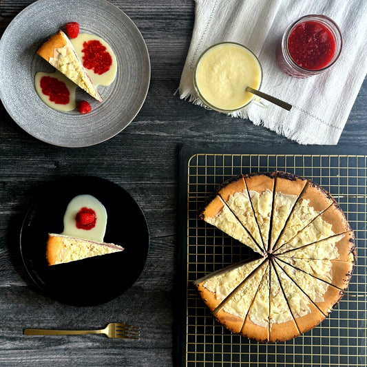 Elegant 10 inch handcrafted white chocolate raspberry cheesecake topped with white chocolate shavings, and two individually plated slices of white chocolate raspberry cheesecake with homemade white chocolate sauce and fresh raspberry garnishes