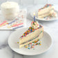 Velvety slice of birthday cake cheesecake with a thick layer of birthday cake baked-in, topped with our sweet cream cheese icing and then covered in colorful sprinkles with an added swirl of homemade whipped icing and a birthday candle