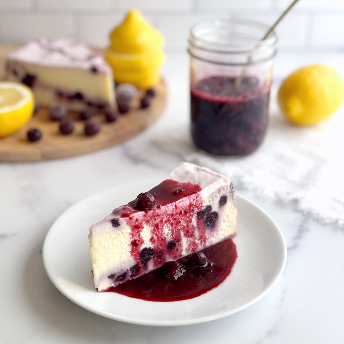Velvety slice of Lemon Blueberry cheesecake with real blueberries baked-in, topped with a sweet blueberry cream cheese icing, and a homemade blueberry compote drizzled on top and dripping from the sides.