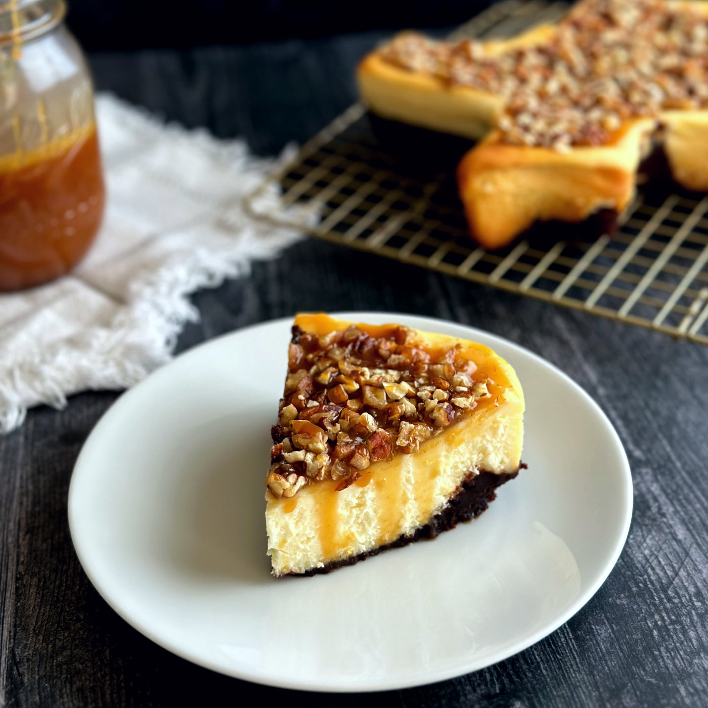 Velvety piece of creamy cheesecake topped with rich caramel and Texas pecans, sitting atop a brownie crust