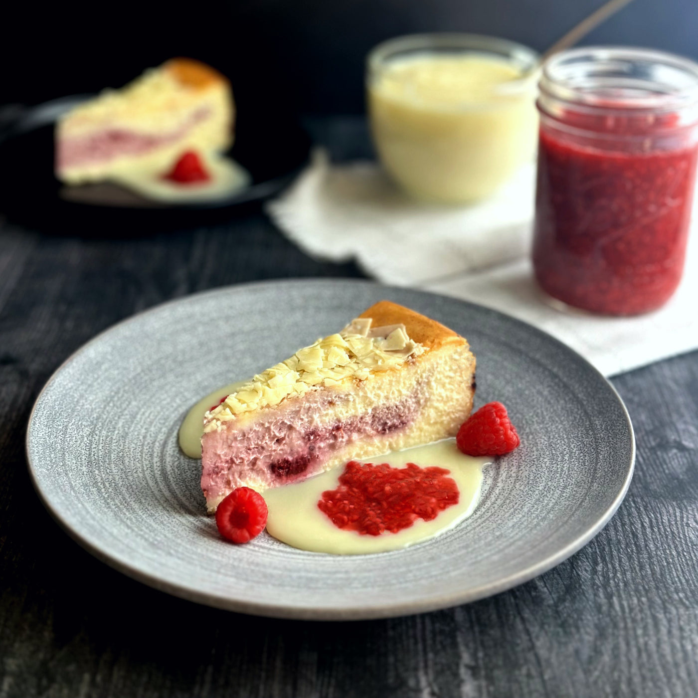 Velvety slice of white chocolate raspberry cheesecake with an elegant layer of handcrafted raspberry filling, topped with white chocolate shavings, all sitting on a delicious puddle of homemade white chocolate and raspberry sauces