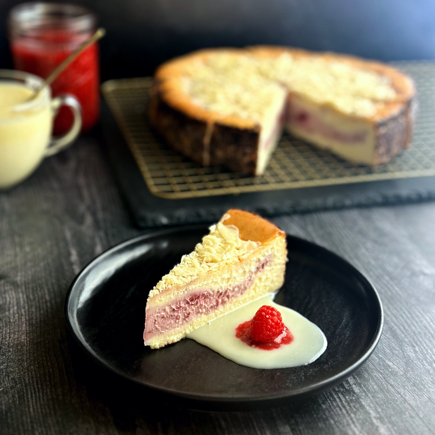 Velvety slice of white chocolate raspberry cheesecake with an elegant layer of handcrafted raspberry filling and topped with elegant white chocolate shavings, with a serving of white chocolate and raspberry sauces on the side, and a fresh raspberry garnish